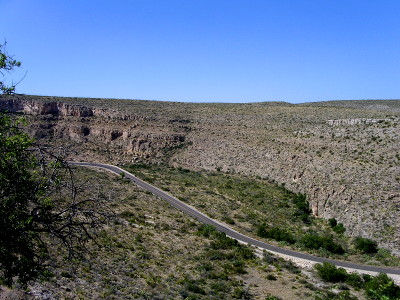 [A paved roadway winds well below the tops of the rocky canyon walls. There is some green vegetation on the valley level where the road is.]